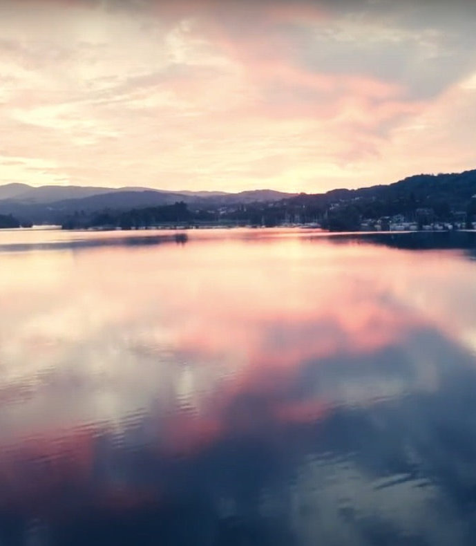 A scenic landscape photograph of a calm lake at sunset, with pink and peach clouds reflected in the water, dark tree-lined shores, and distant mountains.